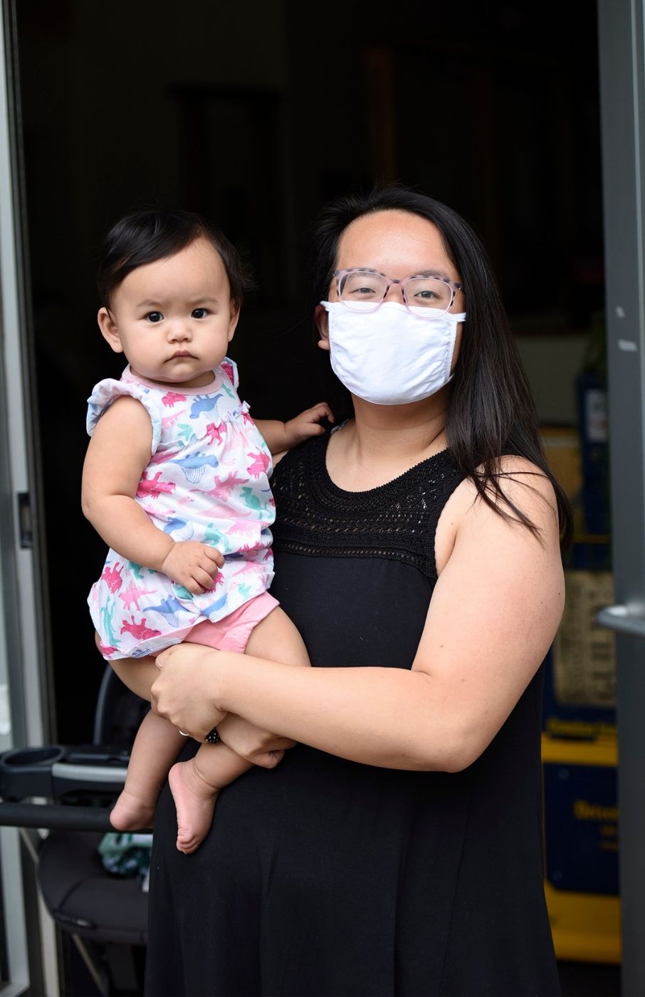 Lily Huang wearing a sleeveless black dress and a white face mask, holding her baby, who's wearing a blue and pink dress. Lily is standing in a doorway. Behind her is a dark room.