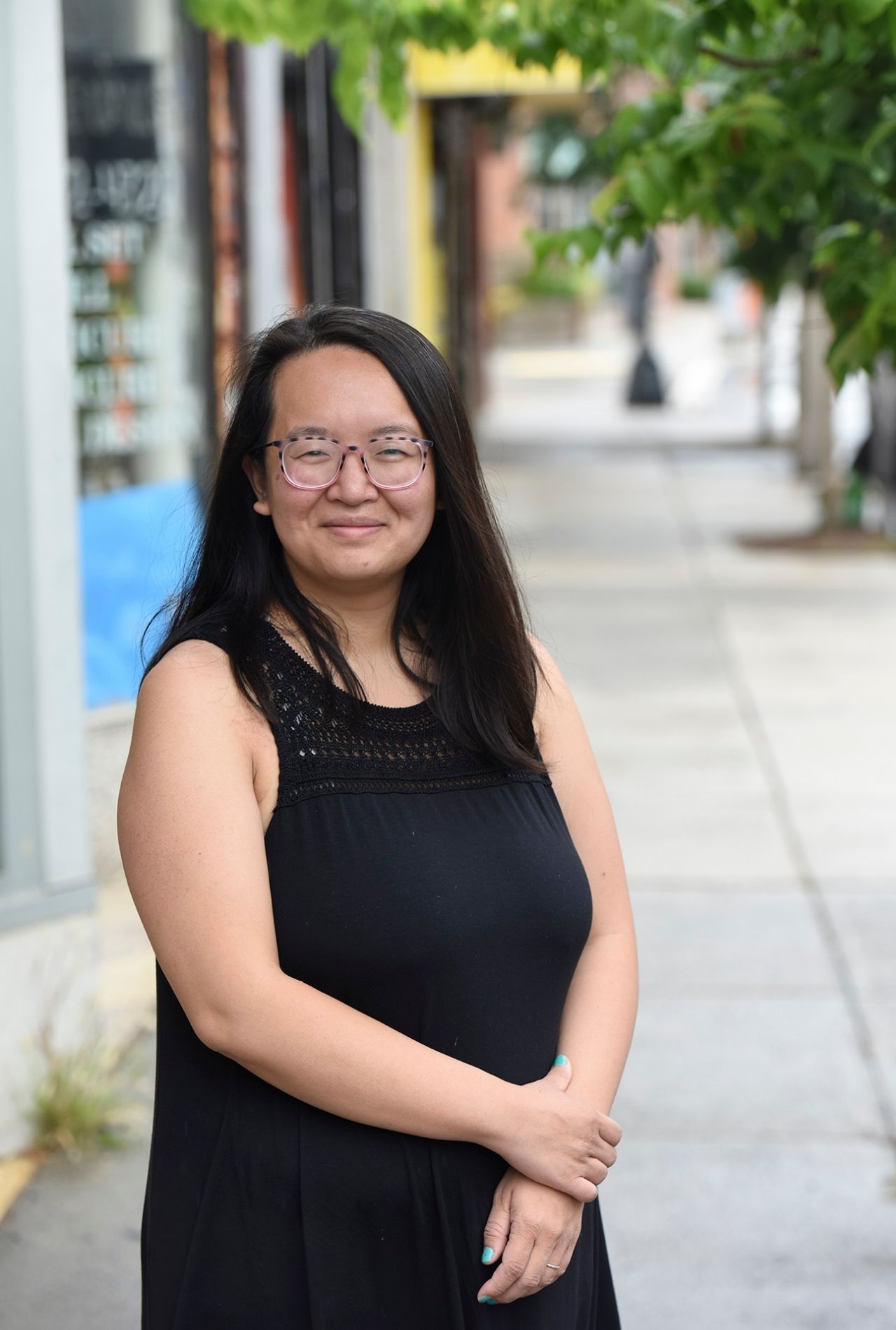 Lily Huang wearing a sleeveless black dress, smiling with her hands folded at her waist, standing on a paved sidewalk with storefronts trailing behind her to the left. Green leaves of a tree peak into the upper right of the image.