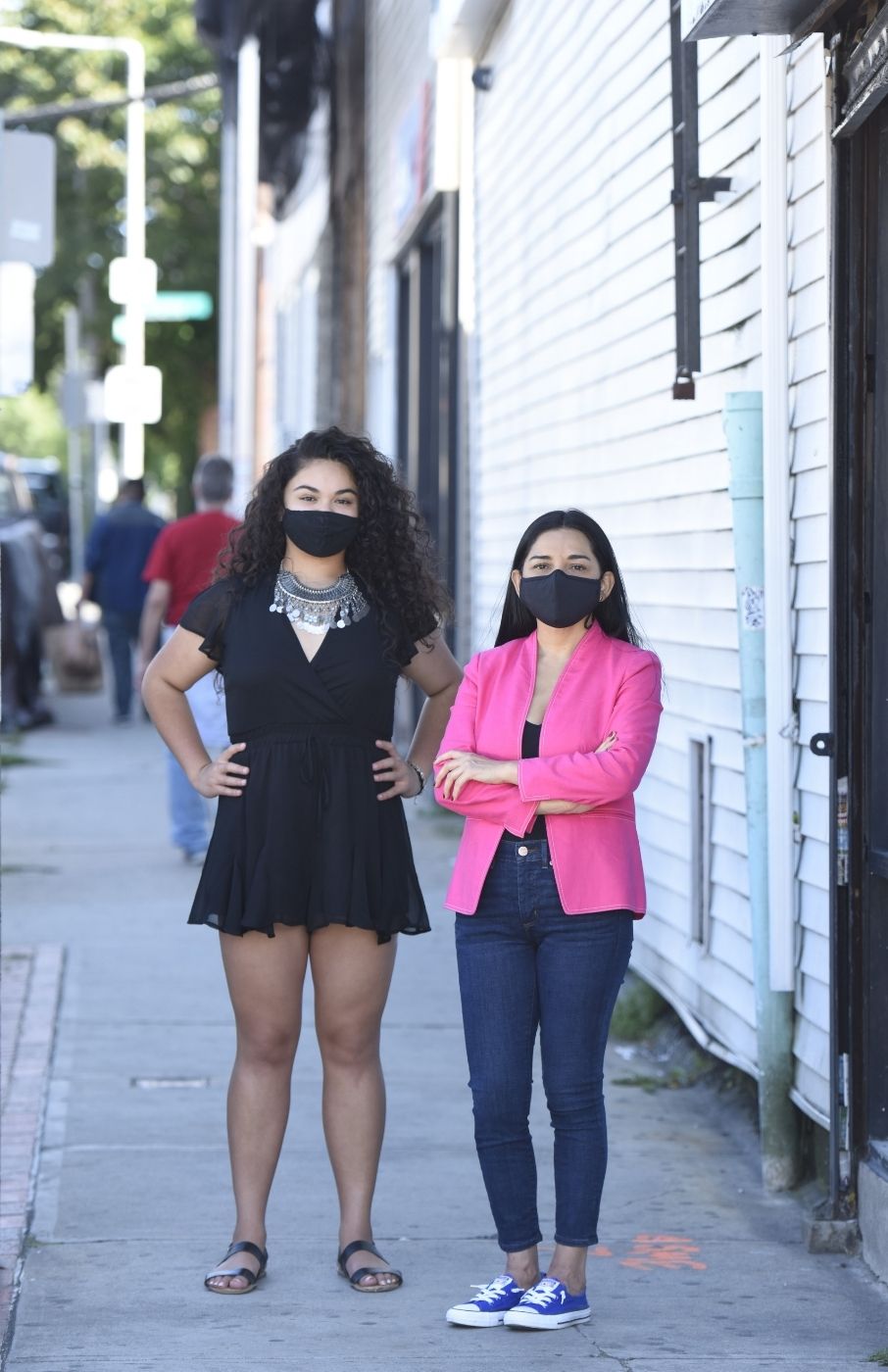 Alexa Cuellar and Patricia Montes standing next to each other. They're both wearing black face masks. Alexa is wearing a short-sleeved black romper and a turquoise necklace, hands on her hips. Patricia is wearing a pink blazer and blue jeans, arms crossed. They're standing on a sidewalk. Behind them to the right is a long white building with siding. Trees and a street sign in the far background.