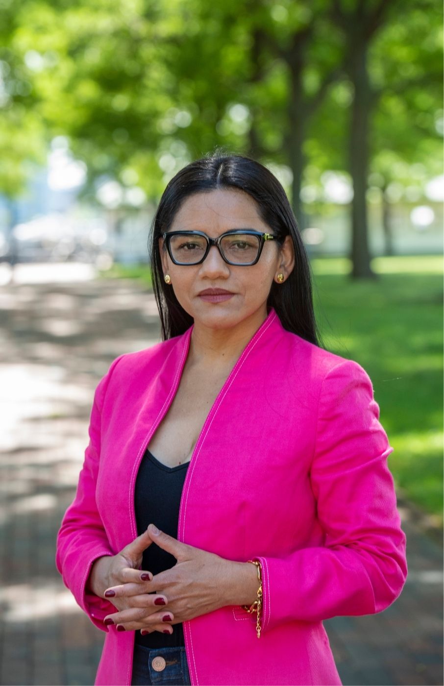 Patricia Montes standing in a park on a sunny day, green trees and grass in background. She's wearing a bright pink blazer, a black shirt and dark blue jeans. She has dark rimmed glasses. She's wearing her long dark hair down.