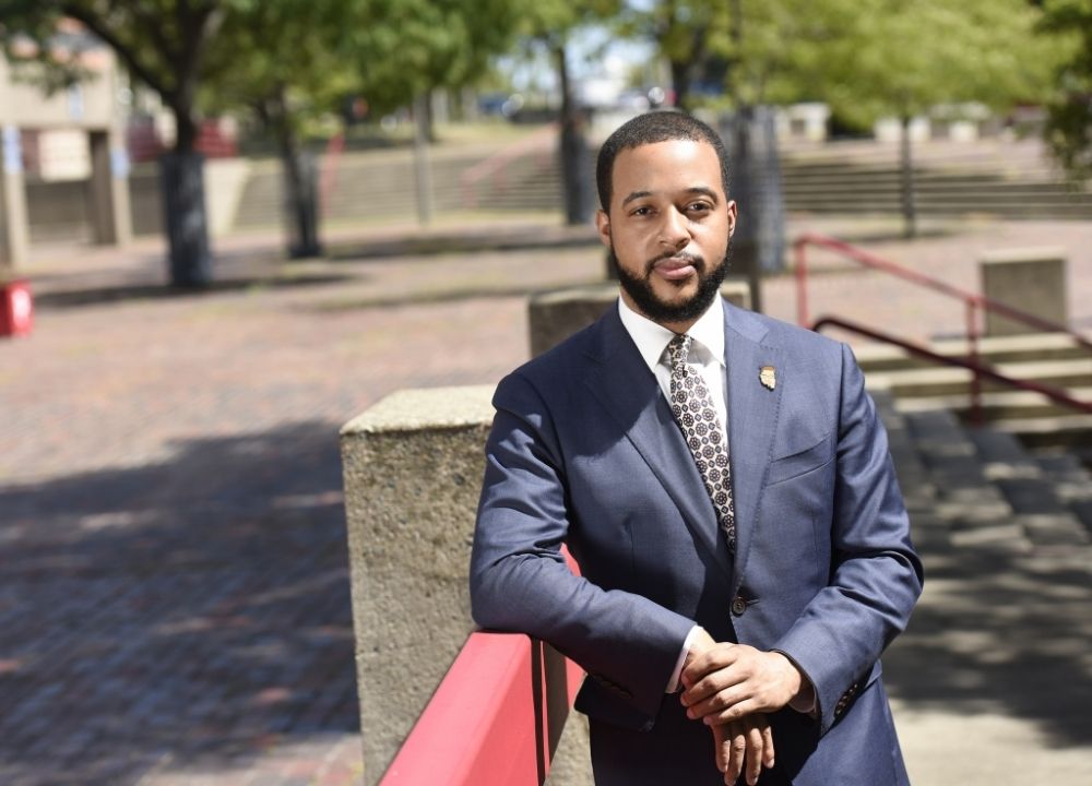 Segun Idowu standing in a park with brick ground. Green trees in the background. He's wearing a dark blue suit, white collared shirt and a red, white and blue patterned tie. He's resting one elbow on a red railing.