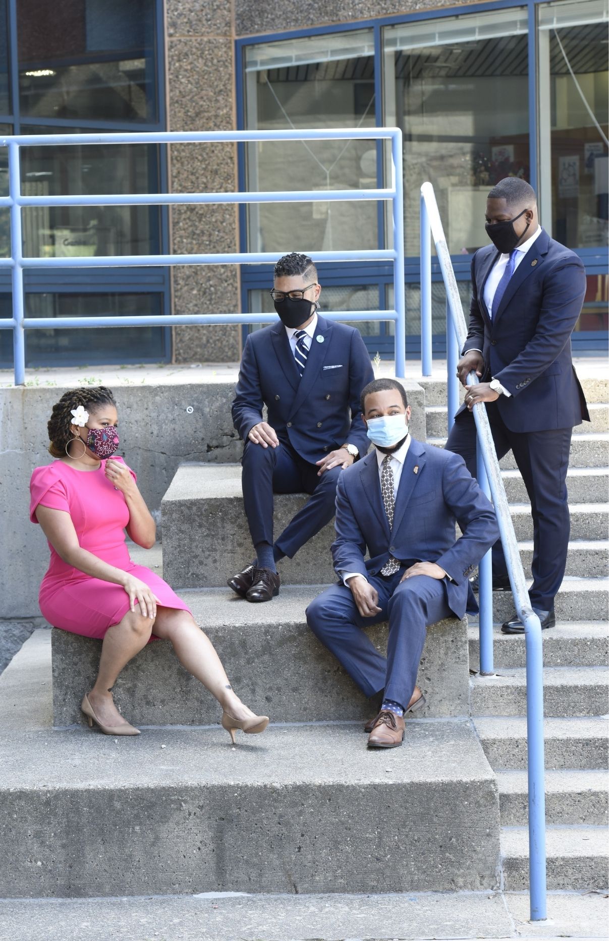 Sheena Collier, Imari Paris Jeffries, Segun Idowu and Willie Bodrick sitting on concrete steps. Everyone but Willie is stilling. Imari, Segun and Willie are wearing dark blue suits. Sheena is wearing a bright pink dress. It looks as though the four of them are having a conversation with one another.