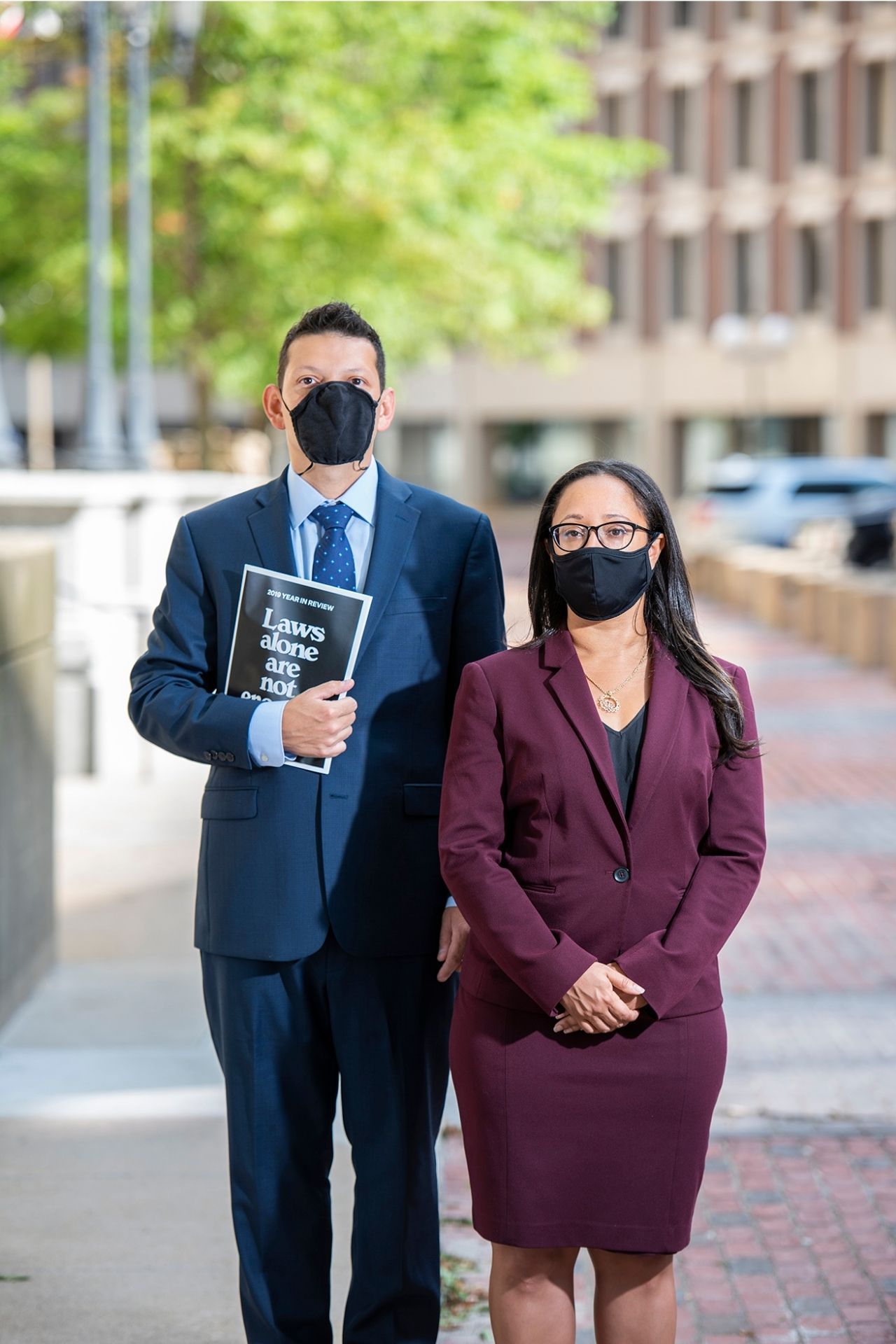 Sophia Hall and Ivan Espinoza Madrigal standing side by side outside in Government Center on a brick walkway. Sophia is wearing a purple blazer and skirt with black shirt. Ivan wearing a dark blue suit and tie and light blue collared shirt. He's holding a black booklet with white text on the cover that says "Laws alone are not enough." Sophia has her hands folded at her waist. They are both wearing black cloth face masks.