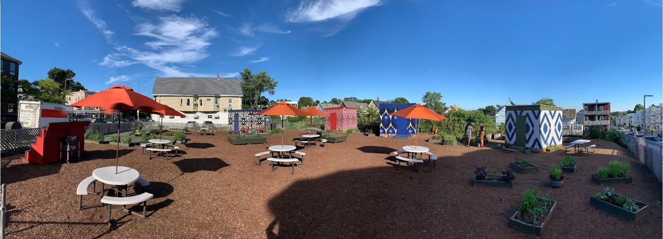 A panoramic shot of The Guild in Dorchester. The sky is blue with few white clouds. white picnic tables with red umbrellas are scattered. The ground is packed first. Colorful, patterned cube structures are in the background.