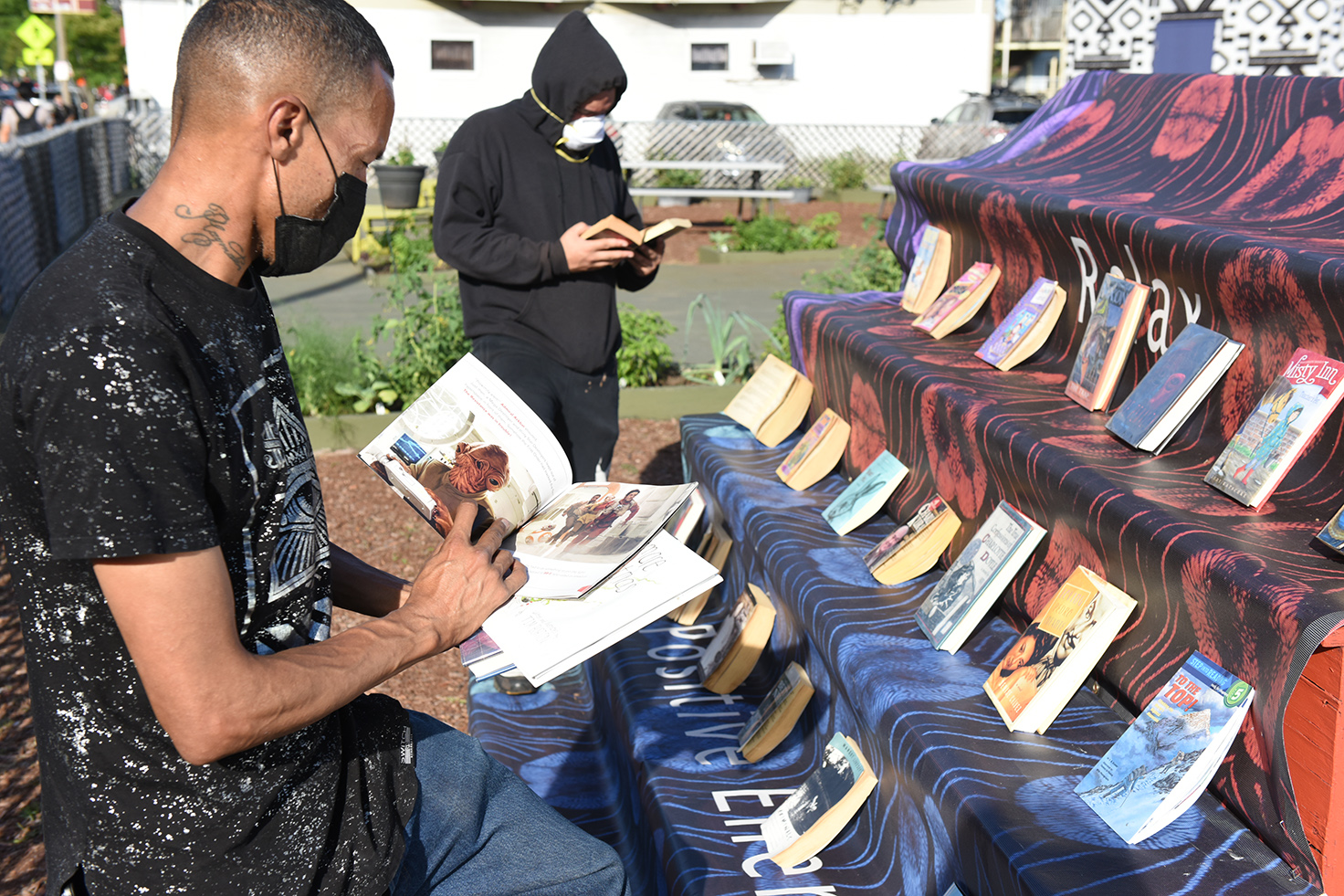 A man standing at an outdoor, tiered bookshelf at The Guild in Dorchester. He's holding a picture book open and reading it. A man in the background does the same. They're both wearing a black t-shirt and black hoodie, respectively. Both are wearing face masks.