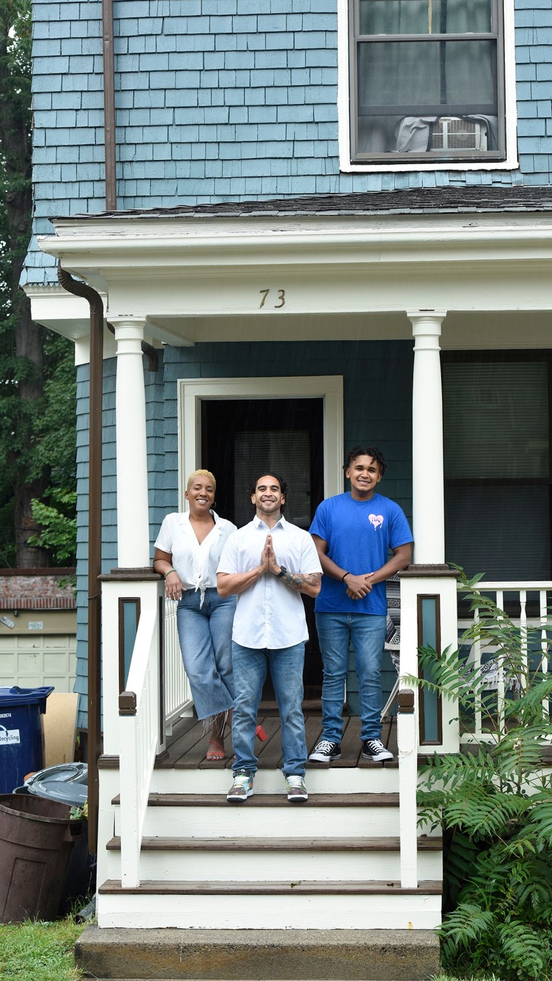 From left to right, Taheera, Mike and Christian Massey standing on the front porch steps of their teal house in Dorchester. The steps are brown and wooden with white molding. Taheera and Mike are wearing jeans a white t-shirts. Christian is wearing a blue T shirt. Green grass a leafy shrubs are in front of/to the right of the steps.