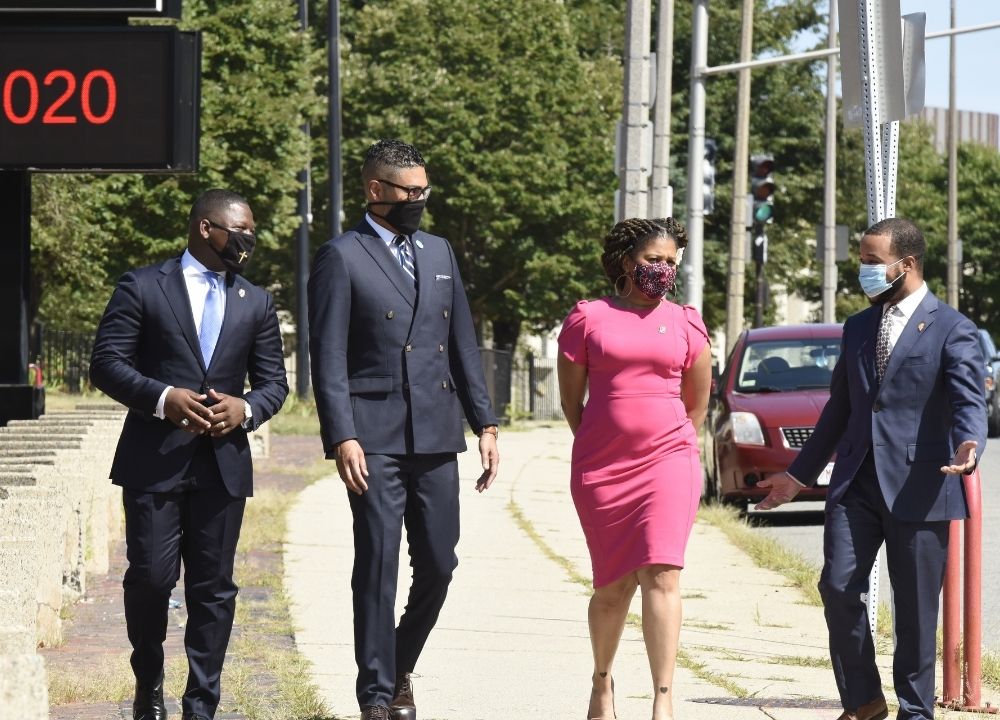 Willie Bodrick, Imari Paris Jeffries, Sheena Collier and Segun Idowu walking next to each other on a sidewalk next to a main road on a sunny day. Willie, Imari and Segun are wearing dark blue suits and Sheena is wearing a bright pink dress. They're all wearing face masks. Green trees are in the background.