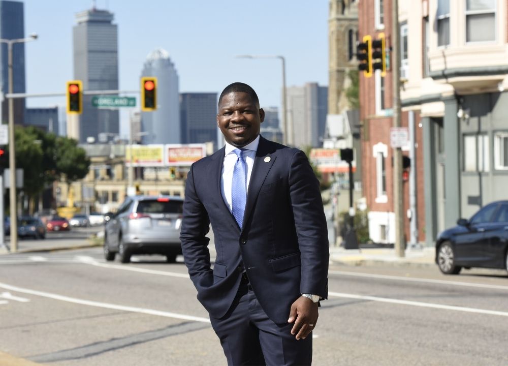 Rev. Willie Bodrick standing center frame on a meridian. In the background is a main road with cars on it, storefronts to the right, and the Prudential Center and One Dalton to the left. The sky is blue. Willie is wearing a dark blue suit, a light blue collared shirt and a blue tie. One hand is in his pocket.