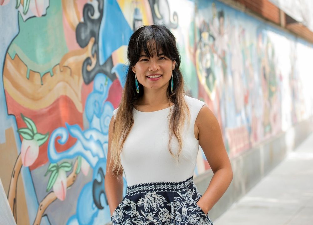 Yanyi Weng is standing on a side walk next to a large, colorful mural to her right that trails behind her. She is smiling, her hands in the pockets of her dress, which is sleeveless, white on the top, and the bottom is an intricate, black and white pattern. She has dark hair and bangs.