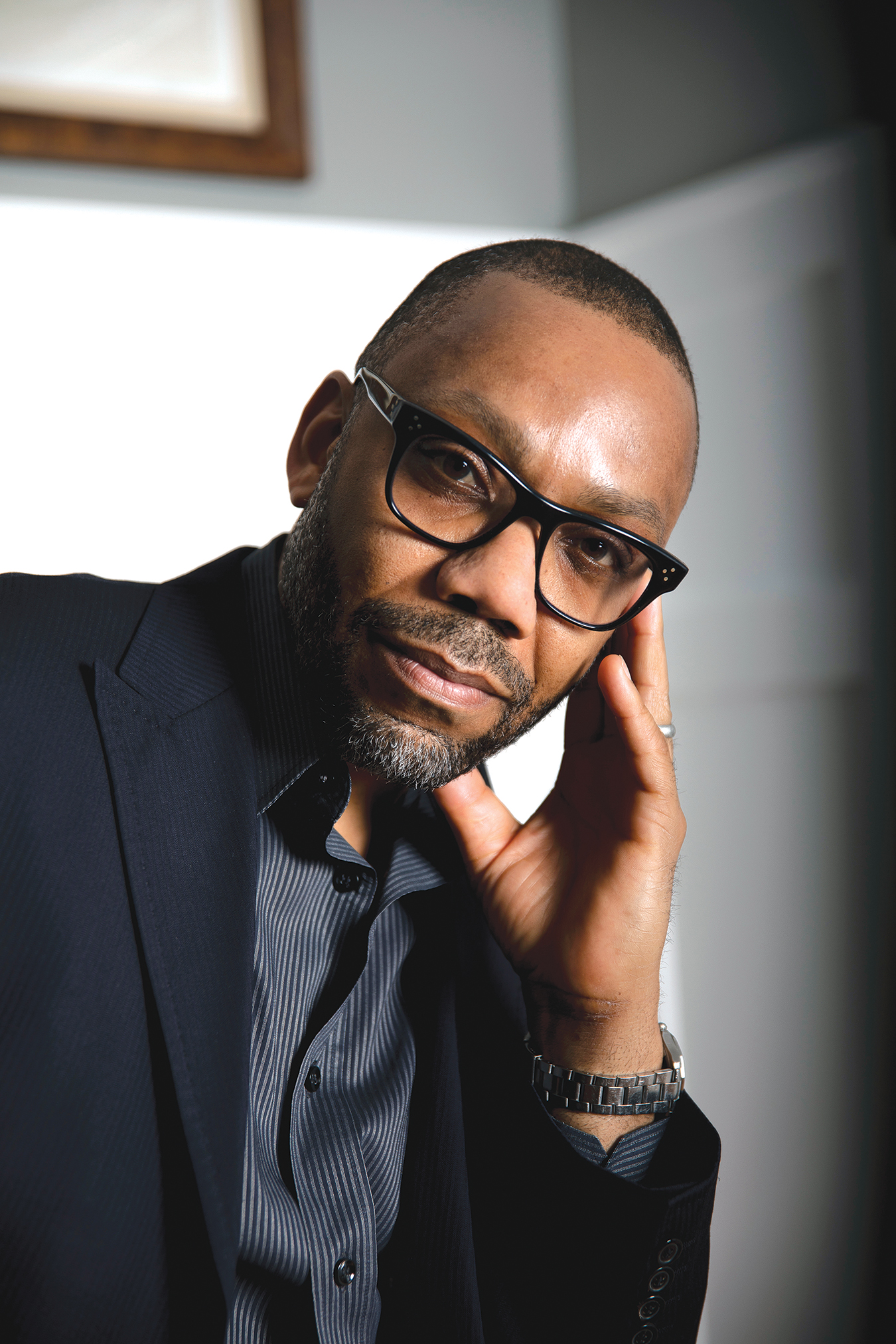 Headshot of Andre Perry. He's leaning his head on his hand, wearing a black blazer and black framed glasses.