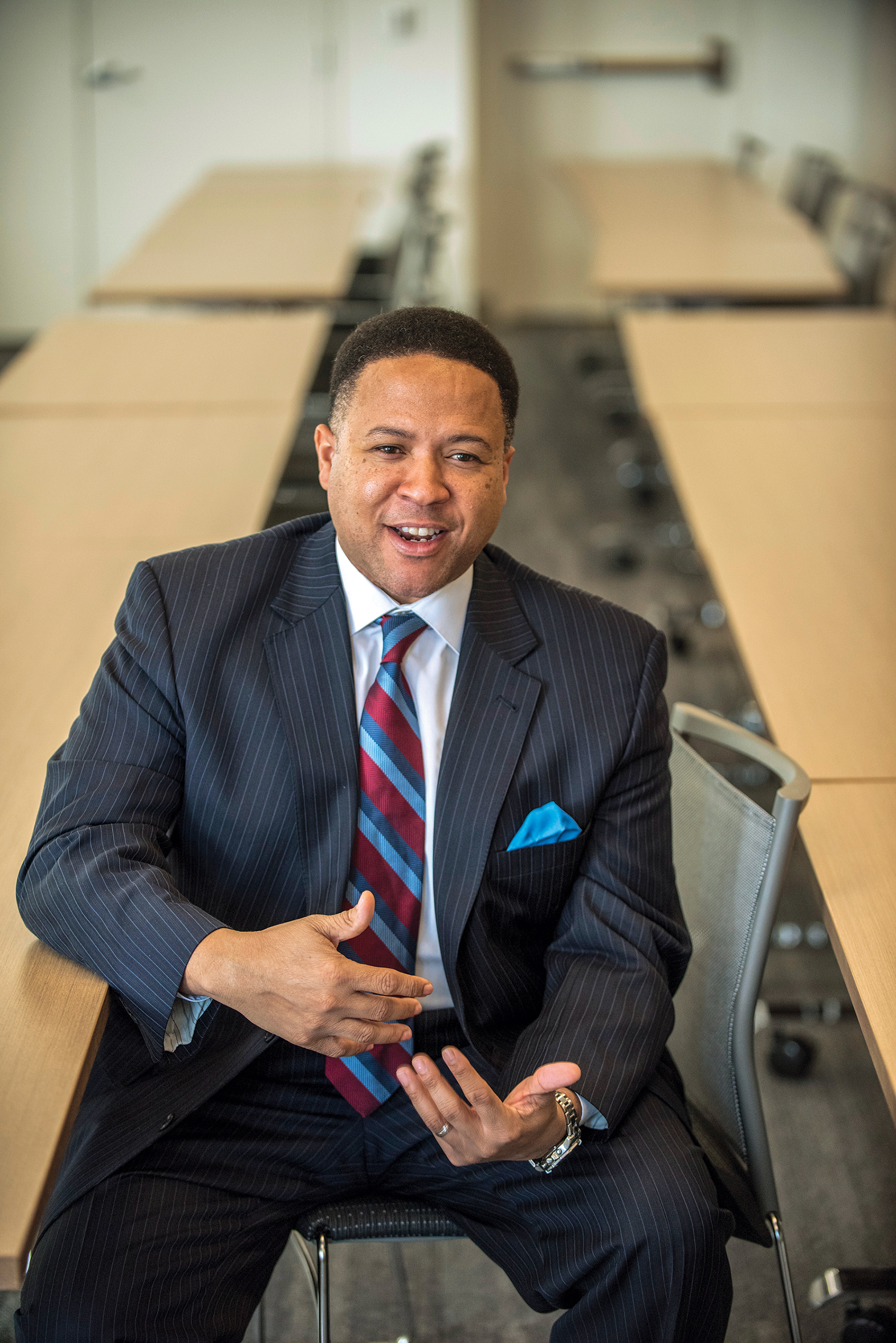 Orlando Wakins, smiling, hands in motion, sitting in a chair with many long tables behind him.