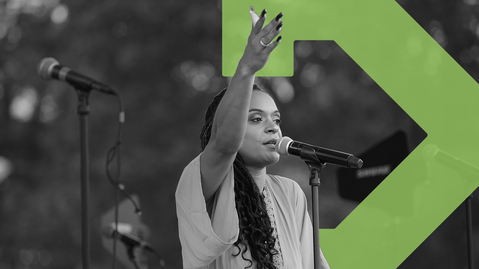 A woman stands onstage speaking into a microphone, she has one arm raised out. Behind the woman is the green TBF arrow.