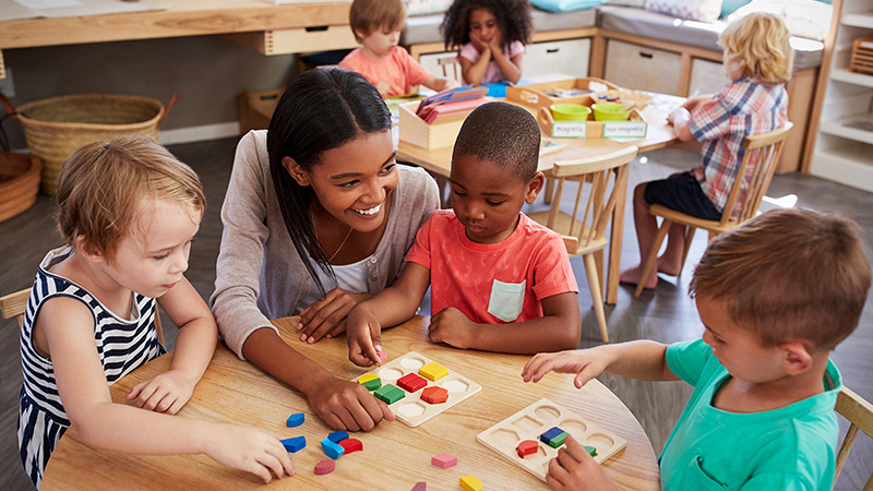 A woman crouches down to a table with three young children seated around it, they are playing a game with colorful block shapes.